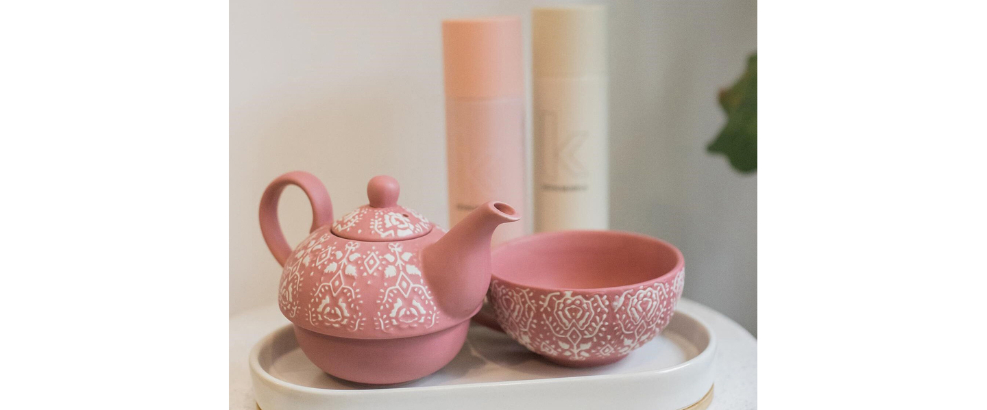 A pink teapot with white decorative patterns and matching cup on a tray beside hair care products, symbolizing green tea benefits for hair.